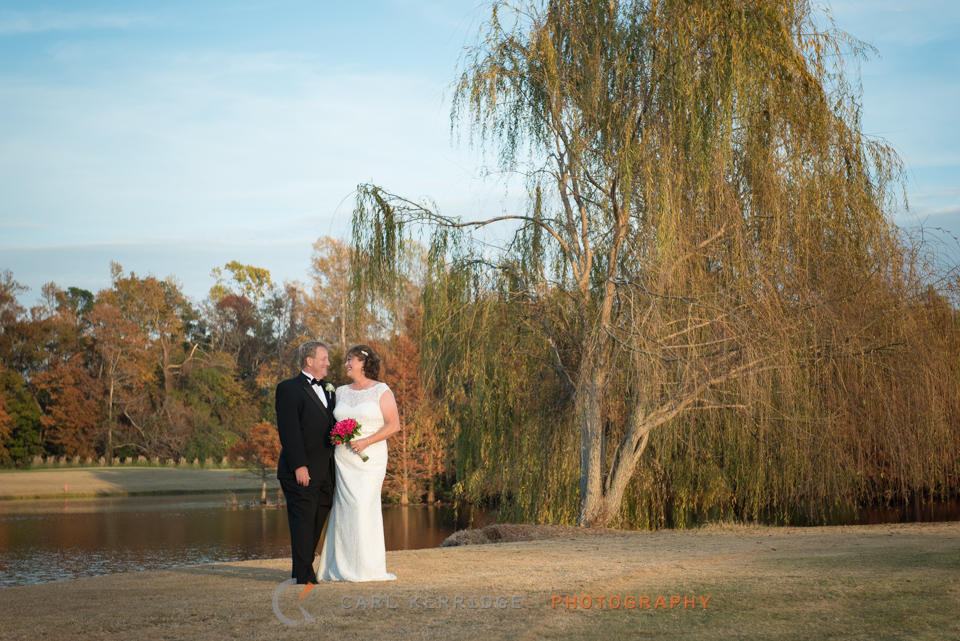 The bride and groom look at each other with love as they pose for a portrait near a willow tree and pond at the members club at grande dunes. 