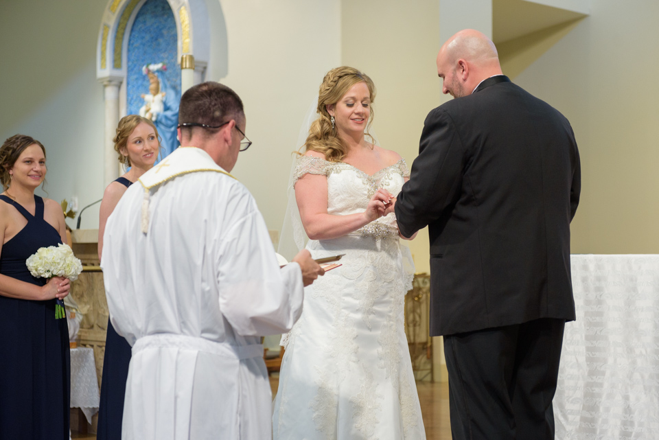 bride places ring on grooms finger in front of the catholic priest